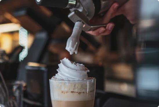 A cream charger/ dispenser being used to add whipped cream to a hot chocolate